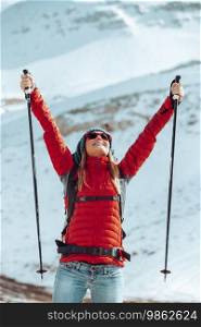 Pretty girl with raised up hands standing among beautiful snowy mountains with walking poles in hands. Looking in the sky. Enjoying winter holidays.. Happy Woman Enjoying Winter Holidays