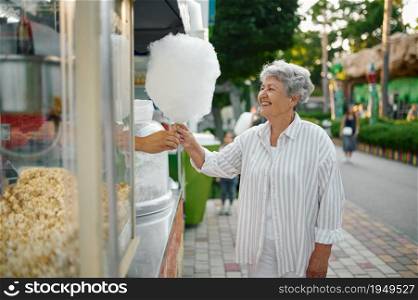 Pretty granny buying cotton candy in summer amusement park. Aged people lifestyle. Funny grandmother having fun outdoors, old female person on nature. Pretty granny buying cotton candy, amusement park