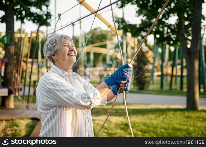 Pretty granny climbing on ropes in summer park. Aged people lifestyle. Funny grandmother having fun outdoors, old female person on nature. Pretty granny climbing on ropes in summer park