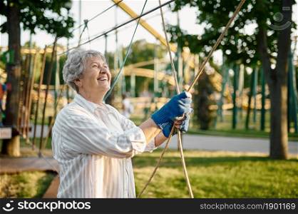 Pretty granny climbing on ropes in summer park. Aged people lifestyle. Funny grandmother having fun outdoors, old female person on nature. Pretty granny climbing on ropes in summer park