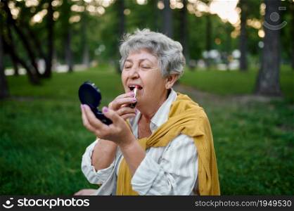 Pretty granny does makeup in summer park, back to youth. Aged people lifestyle. Funny grandmother having fun outdoors, old female person on nature. Pretty granny does makeup in park, back to yout