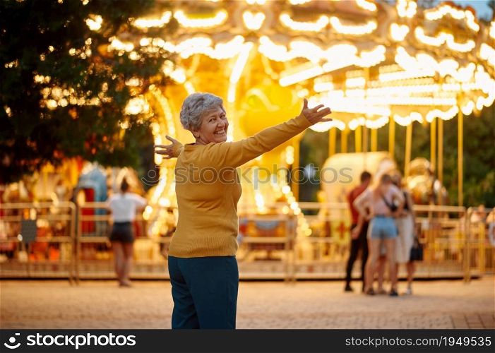 Pretty granny poses in summer amusement park, bright illumination on background, back to childhood. Aged people lifestyle. Funny grandmother having fun outdoors, old female person on nature. Pretty granny, amusement park, bright illumination