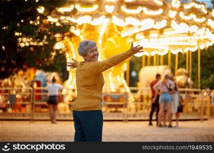 Pretty granny poses in summer amusement park, bright illumination on background, back to childhood. Aged people lifestyle. Funny grandmother having fun outdoors, old female person on nature. Pretty granny, amusement park, bright illumination
