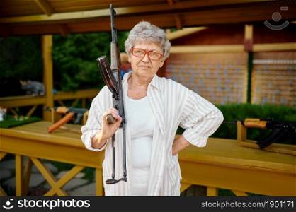 Pretty granny with gun poses in shooting gallery, amusement park. Aged people lifestyle. Funny grandmother having fun outdoors, old female person on nature. Pretty granny with gun poses in shooting gallery