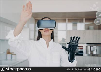 Pretty handicapped girl in virtual reality glasses at home. Disabled person gets rehabilitation and treatment. Young caucasian woman touches 3d cyber vision. Amputee has modern bionic limb.. Disabled girl with modern bionic limb in virtual reality glasses. 3d vision for rehabilitation.