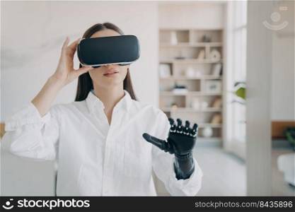Pretty handicapped girl in vr glasses at home. Disabled person gets rehabilitation. Young european woman in white with artificial hand excited with 3d cyber vision. Futuristic medical technology.. Pretty handicapped girl in vr glasses at home. Futuristic medical technology for rehabilitation.