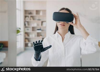 Pretty handicapped girl in vr glasses at home. Disabled person gets rehabilitation. Young european woman in white with artificial hand excited with 3d cyber vision. Futuristic medical technology.. Pretty handicapped girl in vr glasses at home. Futuristic medical technology for rehabilitation.