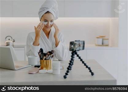 Pretty happy female blogger records streaming video for her blog gives tutorial how to apply beauty patches gives advice about skin care dressed in bathrobe looks at camera poses at table in kitchen