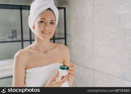 Pretty hispanic woman holding mockup cosmetic jar with cream or serum, advertising skincare moisturizing beauty product. Woman in towel after shower shows template cosmetics package in bathroom.. Woman holding mockup cosmetics jar with cream, advertising skincare beauty product in bathroom