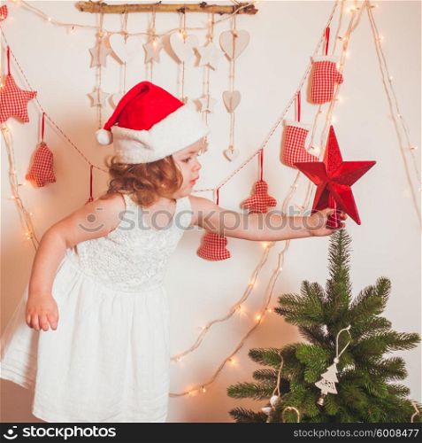 Pretty little girl in a cap of Santa Claus hangs up a star on top of Christmas tree. Christmas time mood