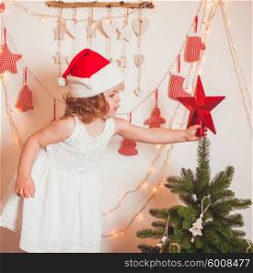 Pretty little girl in a cap of Santa Claus hangs up a star on top of Christmas tree. Christmas time mood