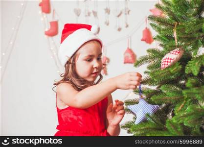 Pretty little girl in a cap of Santa Claus is decorating Christmas tree. Christmas time mood