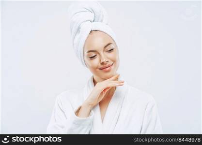 Pretty natural woman touches gently jawline, wears bath towel wrapped on head, dries hair after shower, cares about body, has clean skin, prepares for evening party, dressed in domestic robe