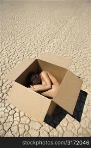 Pretty nude young woman curled up in fetal position inside box in cracked desert landscape in California.