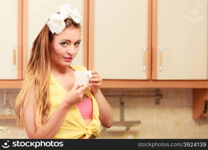 Pretty pin up girl with hairband bow drinking tea or coffee at home. Gorgeous young retro woman with hot beverage relaxing in kitchen.