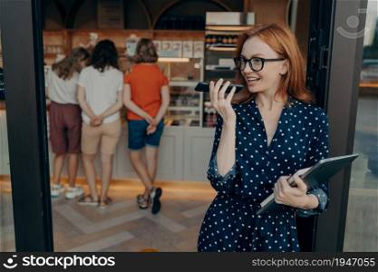 Pretty red haired woman records audio message via cellphone carries laptop and diary looks away poses against shop background indoor wears polka dotted dress. People and technologies concept. Woman records audio message via cellphone carries laptop and diary looks away