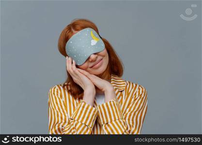 Pretty redhead woman wears blindfold on eyes, keeps hands pressed together near face, sees pleasant dreams while sleeping, dressed in domestic clothes, isolated on grey wall, feels comfortable