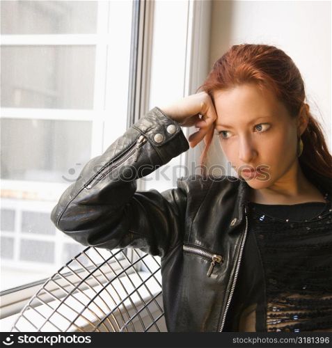 Pretty redhead young woman sitting indoors on metal chair looking out window.