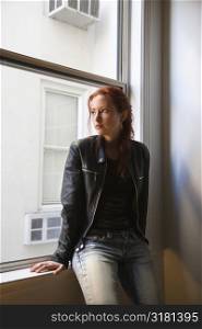 Pretty redhead young woman sitting indoors on window ledge looking out.