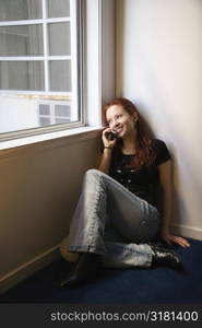 Pretty redhead young woman sitting on floor indoors by window holding cellphone to ear smiling.