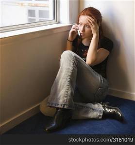 Pretty redhead young woman sitting on floor indoors by window talking on cellphone.