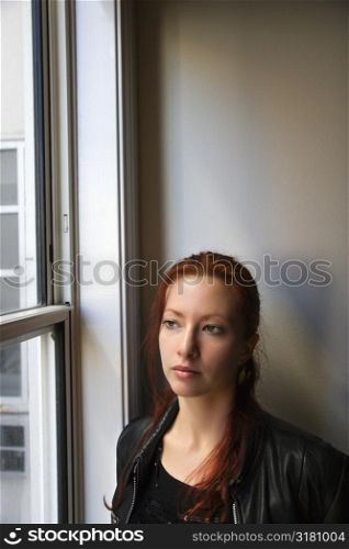 Pretty redhead young woman standing indoors by window looking out.