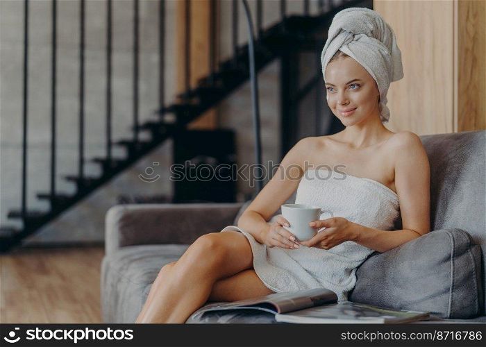 Pretty relaxed young woman looks thoughtfully into distance, holds cup of hot beverage, rests after showering, wrapped in bath towel, sits on couch, reads magazine, poses against cozy interior