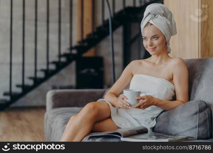 Pretty relaxed young woman looks thoughtfully into distance, holds cup of hot beverage, rests after showering, wrapped in bath towel, sits on couch, reads magazine, poses against cozy interior