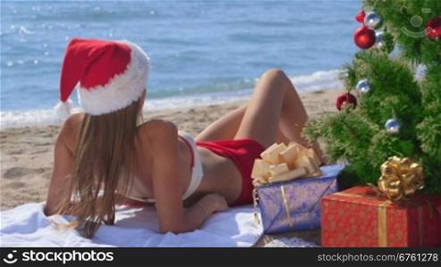 Pretty Santa with gift boxes under Christmas tree on a sandy beach enjoying vacation