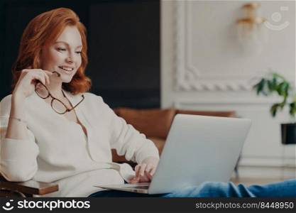 Pretty satisfied redhead female student watches tutorial video, works distantly from home, concentrated in laptop computer, prepares course work, holds spectacles, poses in modern spacious room