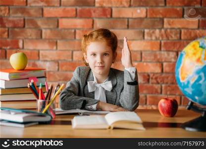 Pretty schoolgirl with raised hand sitting at the table with textbooks, apples and globe. Female pupil at the desk wants to answer