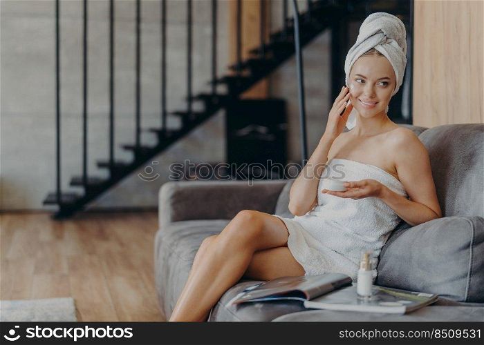 Pretty smiling European woman applies face cream on face, holds jar of cosmetic product, takes care of skin and complexion, poses on comfortable sofa with magazine against stairs background.