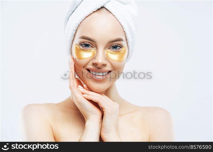 Pretty smiling lady has towel on head touches cheeks, applies golden hydrogel patches, stands shirtless indoor, removes wrinkles, isolated on white wall, has healthy fresh facial skin. High resolution