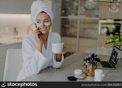 Pretty smiling woman applies hydrogel patches under eyes enjoys grooming herself after shower dressed in bathrobe drinks tea has telephone conversation sits in chair at home. Face care procedures