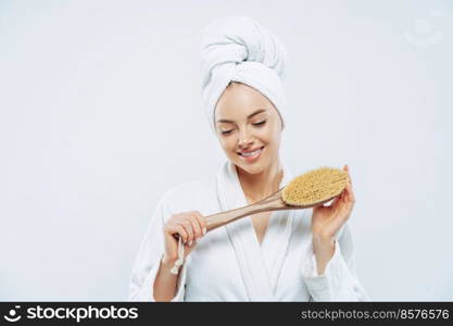 Pretty smiling woman concentrated down, holds bath brush, takes shower, cares about hygiene, wears towel on head, white soft robe, poses against white background. Women and cleanliness concept