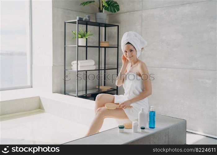 Pretty smiling woman sitting on bathtub during spa skincare treatment procedure. Female holding massaging brush posing in modern bathroom. Body care, natural cosmetics advertising.. Pretty woman enjoy spa skincare treatment procedure in modern bathroom. Body care cosmetics ad