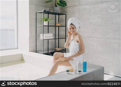 Pretty smiling woman sitting on bathtub during spa skincare treatment procedure. Female holding massaging brush posing in modern bathroom. Body care, natural cosmetics advertising.. Pretty woman enjoy spa skincare treatment procedure in modern bathroom. Body care cosmetics ad