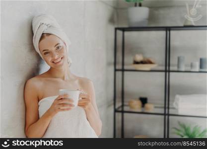 Pretty smiling woman with healthy skin wrapped in white towel, stands glad indoor, drinks coffee, has positive face expression, poses indoor. People, spa, p&ering, skin care and wellness concept