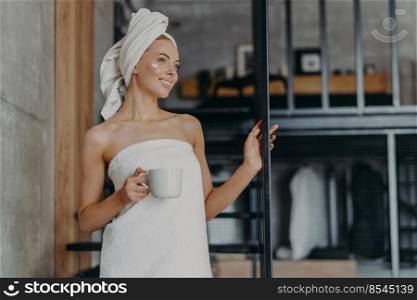 Pretty thoughtful woman concentrated somewhere, smiles broadly, has white perfect teeth, applies face cream, stands wrapped in towel, drinks hot tea, stands on stairs indoor, relaxes after taking bath