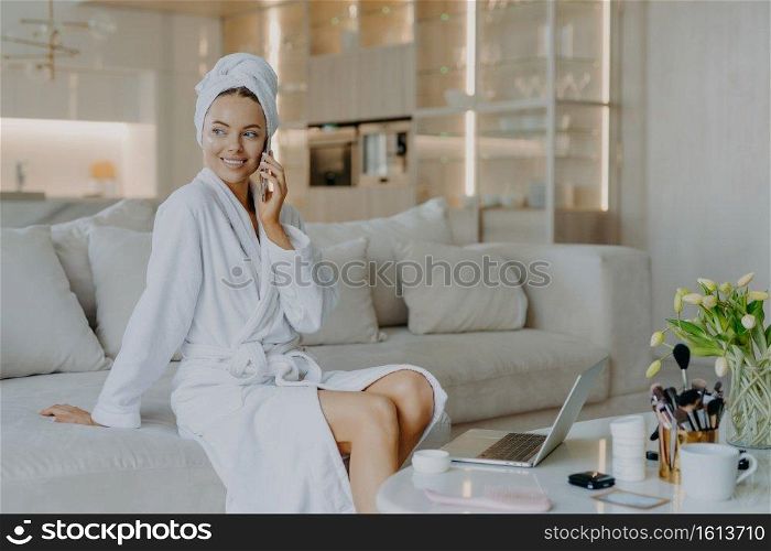 Pretty thoughtful woman with healthy skin well cared complexion talks via mobile phone enjoys cozy domestic atmosphere looks aside sits on comfortable sofa in living room applies cream on face
