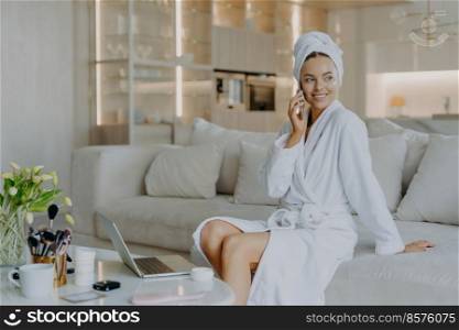 Pretty thoughtful woman with healthy skin well cared complexion talks via mobile phone enjoys cozy domestic atmosphere looks aside sits on comfortable sofa in living room applies cream on face