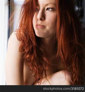 Pretty topless redhead young woman sitting by window looking out.