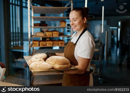 Pretty woman baker holding variety of baked bread on tray standing over bakery house background. Pretty woman baker holding variety of baked bread on tray