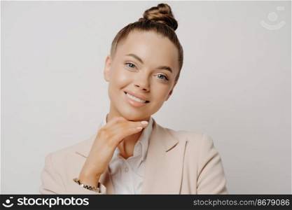 Pretty woman executive in beige suit and hair in bun at business conference listening to good news, being happy and relaxed while looking at camera with hand on her chin isolated on grey background. Woman executive at conference listening to good news