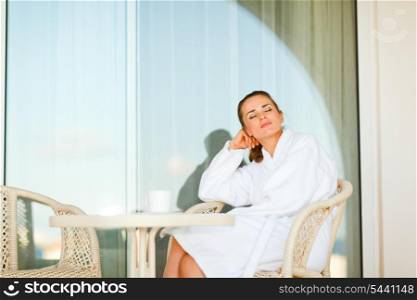 Pretty woman in bathrobe sitting at table on terrace and relaxing&#xA;