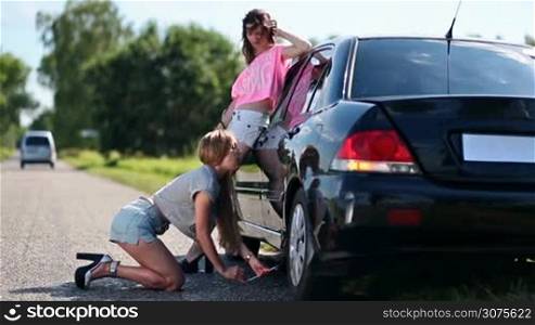 Pretty woman on high heels and shorts is kneeling and jacking up her car to change flat tire with spare one. Her girlfriend is standing near car giving her advice on how to use screw-jack.