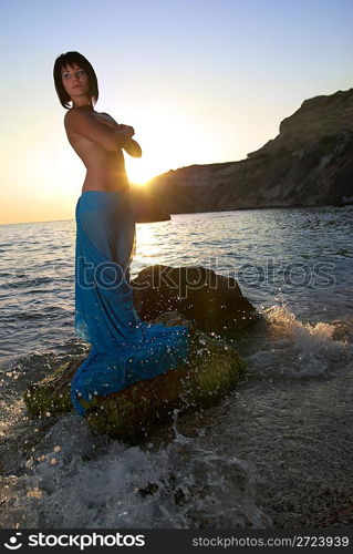 Pretty woman on the sea rock against sunset.