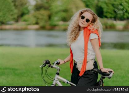 Pretty woman rides bicycle, has bushy blonde hair, stands near her bike, wears sunglasses and white t shirt, enjoys traveling on fresh air, small river or lake in background, green grass. Lifestyle