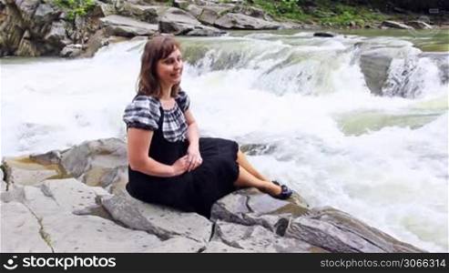 pretty woman sits on stone and looks at mountain river