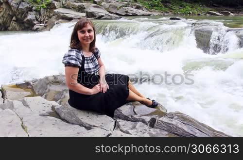 pretty woman sits on stone by shore of mountain river